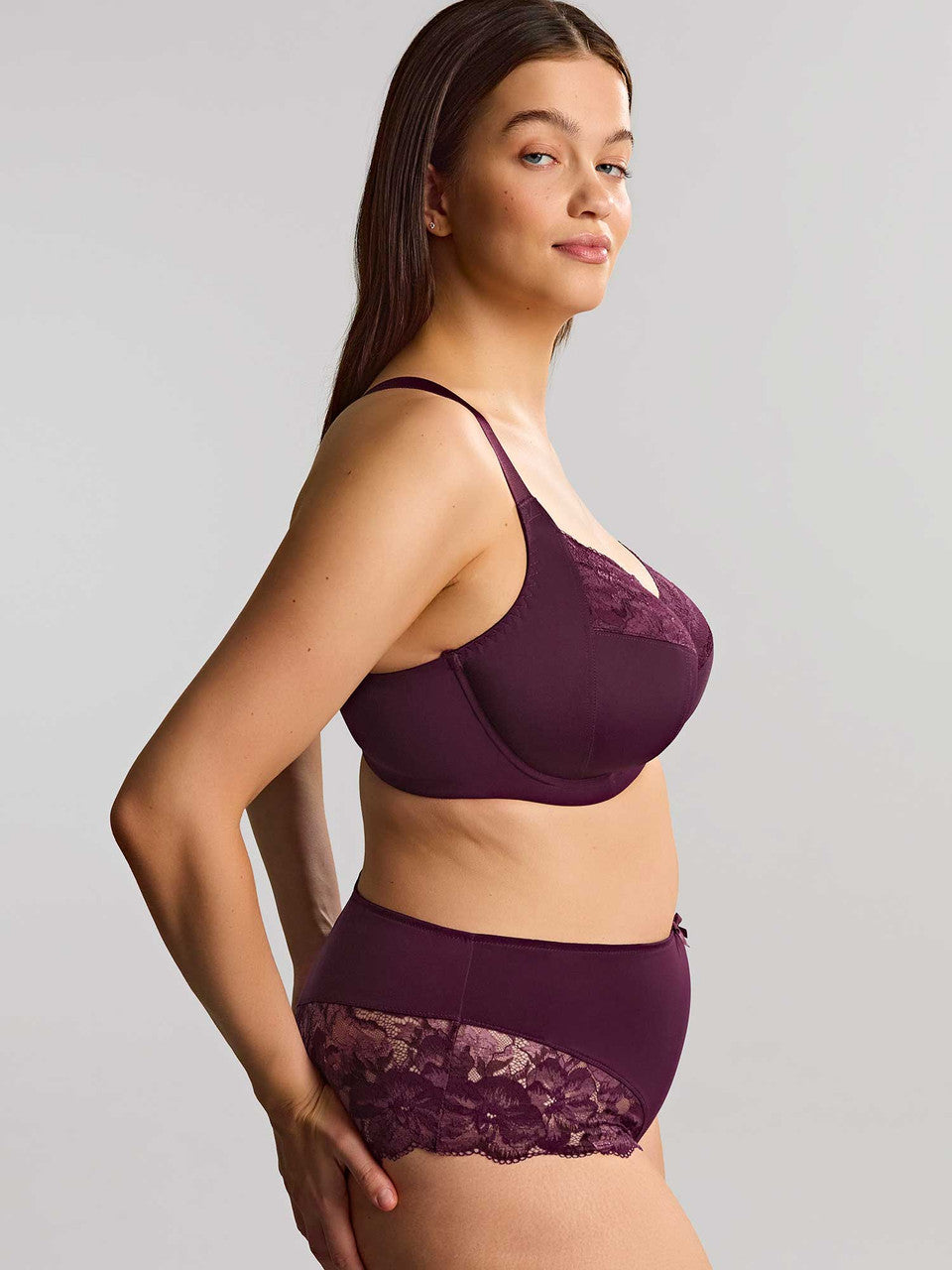 A woman shown from the side wearing a matching lingerie set in deep plum against a gray background.