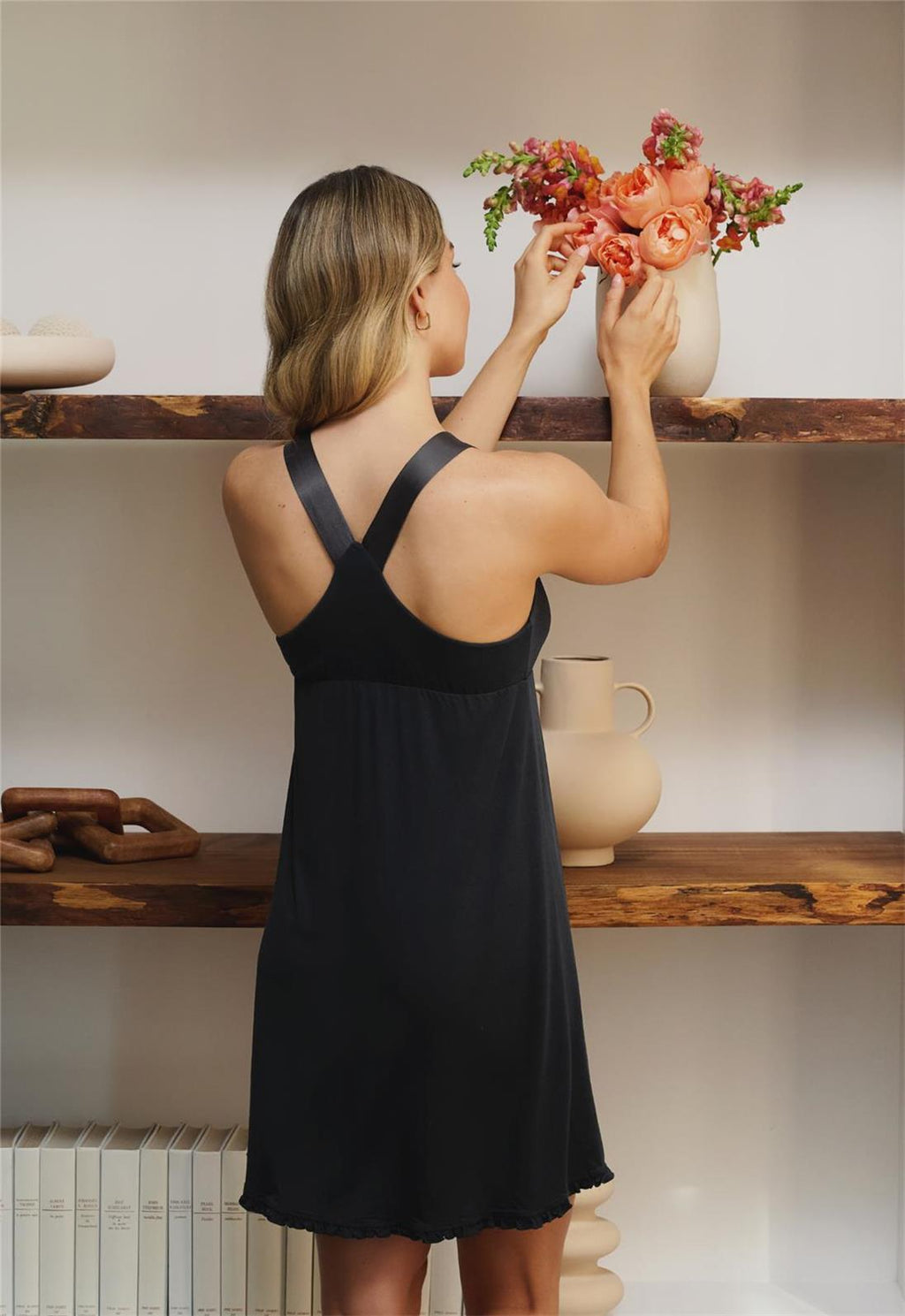 Woman in a black dress arranging flowers in a vase on a wooden shelf.