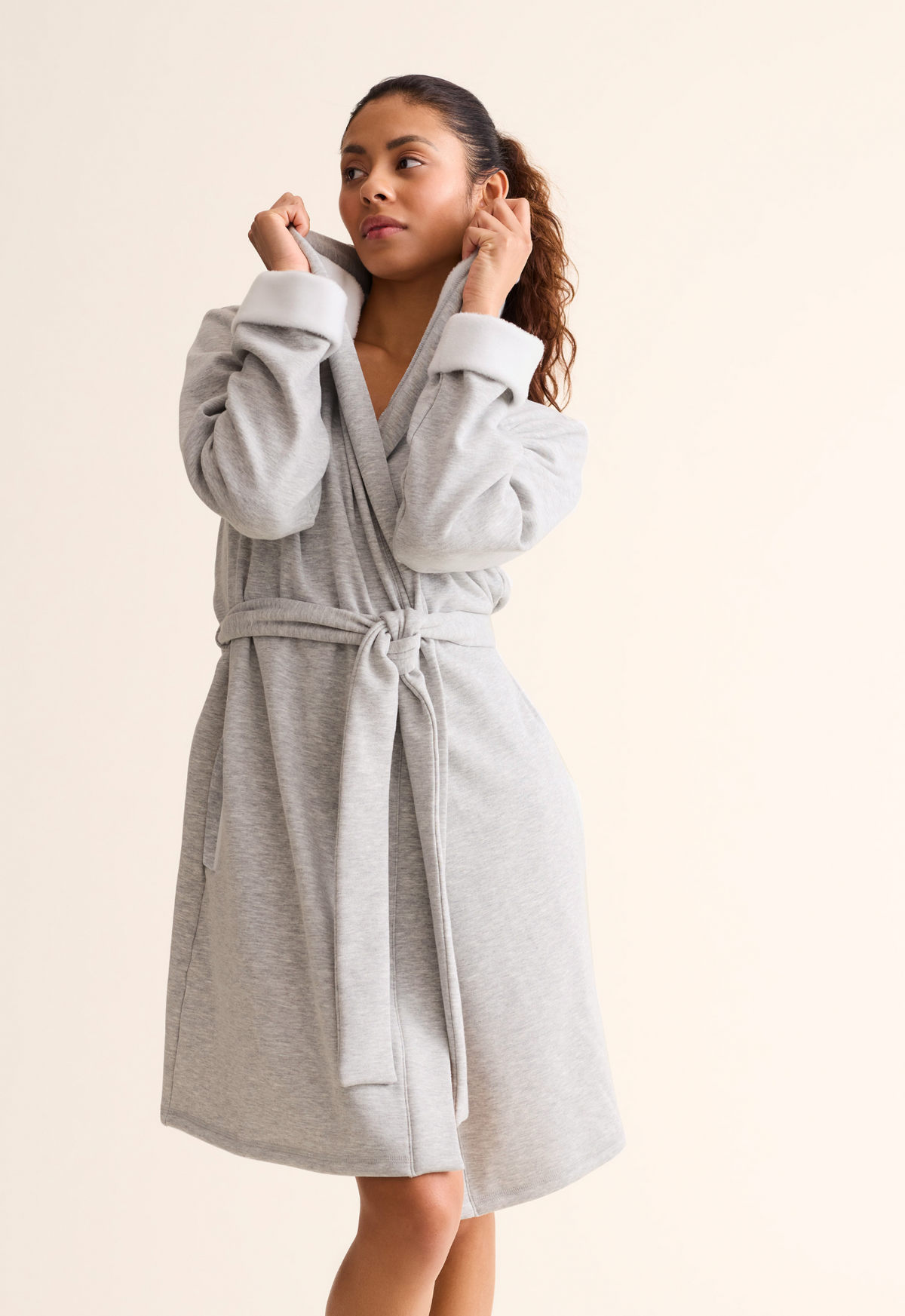 A woman wearing a light gray fleece robe with a tie belt, against a solid light beige background.
