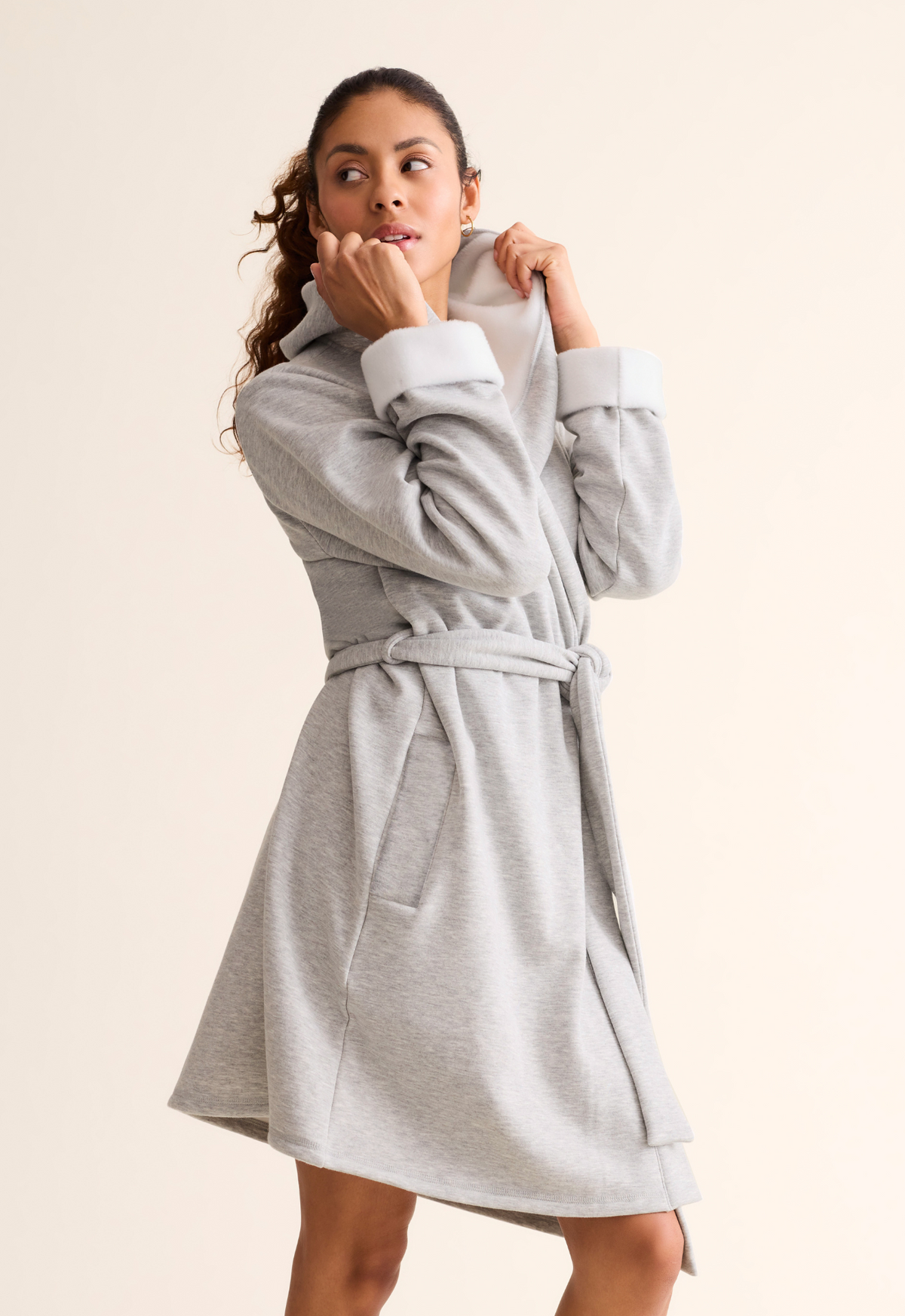 A woman wearing a light gray heathered hooded robe standing against a plain beige studio background.