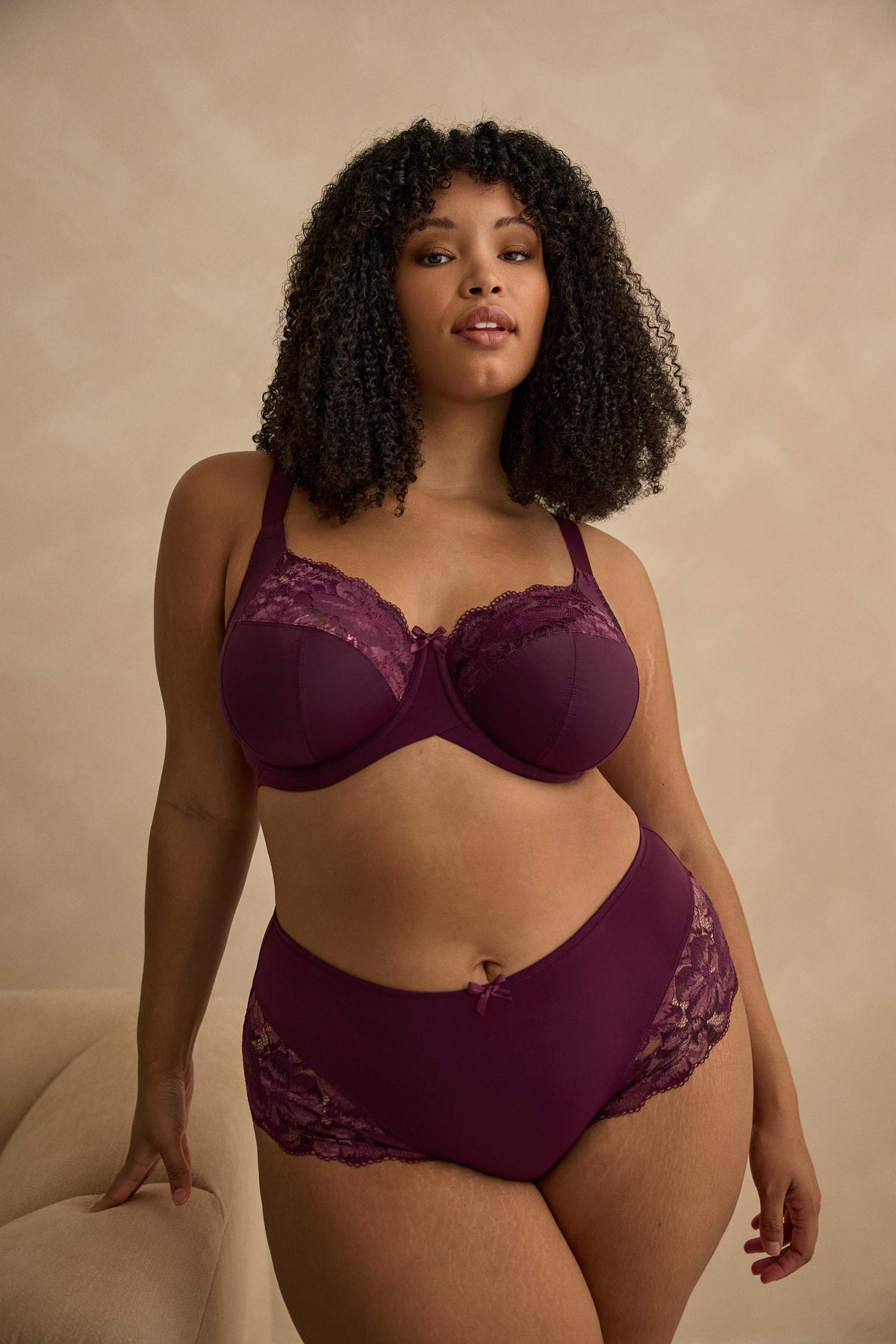 A woman modeling a matching two-piece lingerie set in deep plum purple, against a gray background
