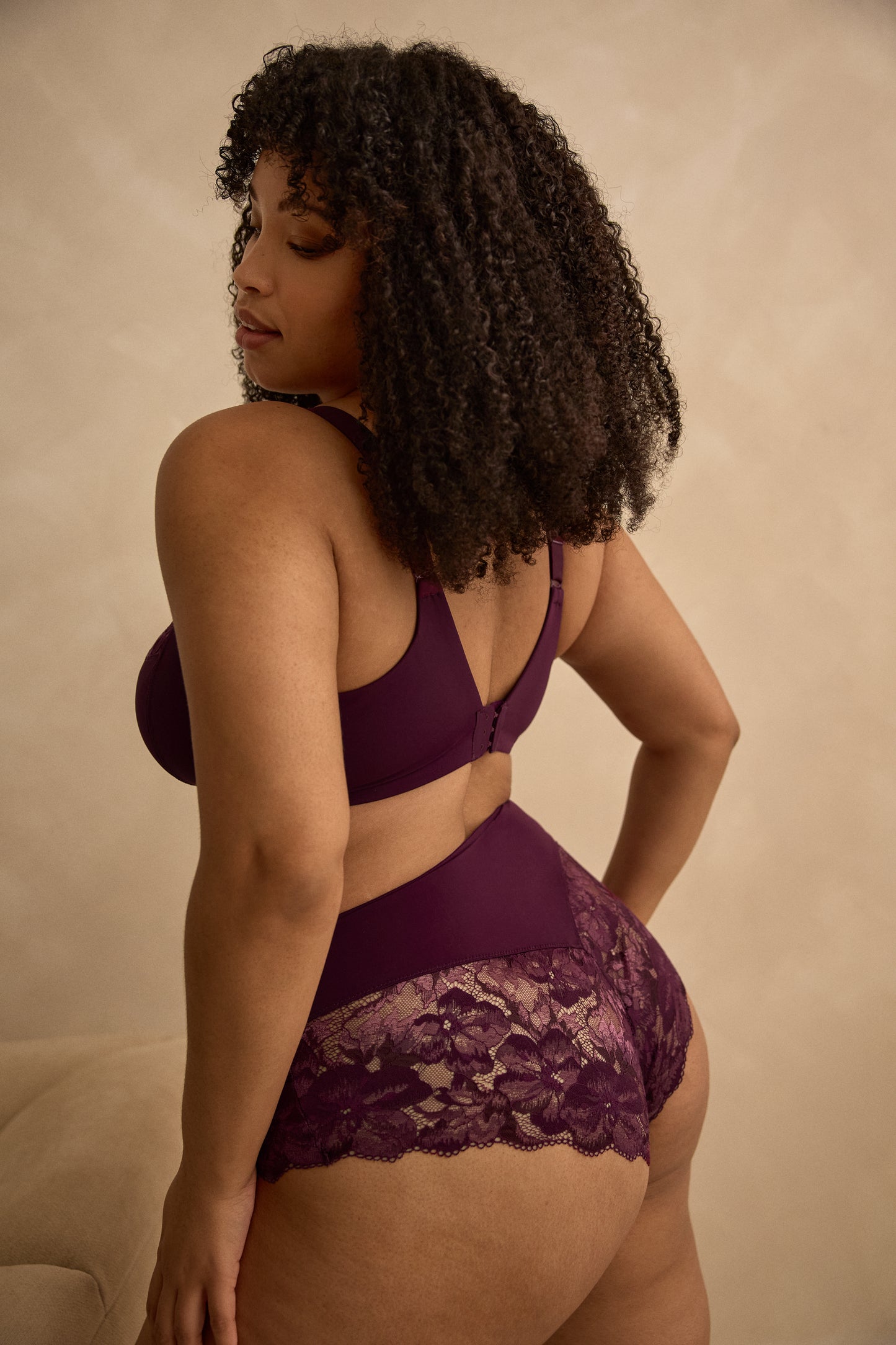 A woman modeling a matching two-piece lingerie set in deep plum purple, against a gray background