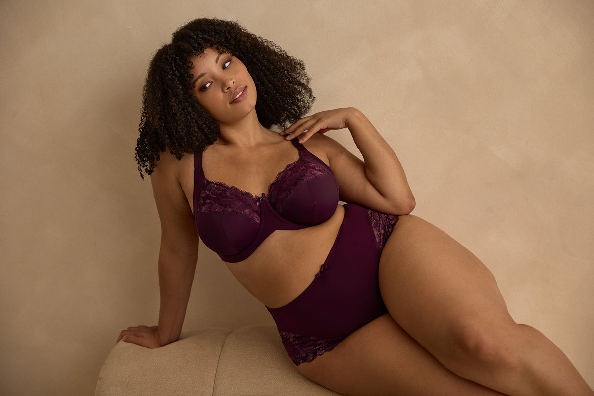 A woman modeling a matching two-piece lingerie set in deep plum purple, against a gray background