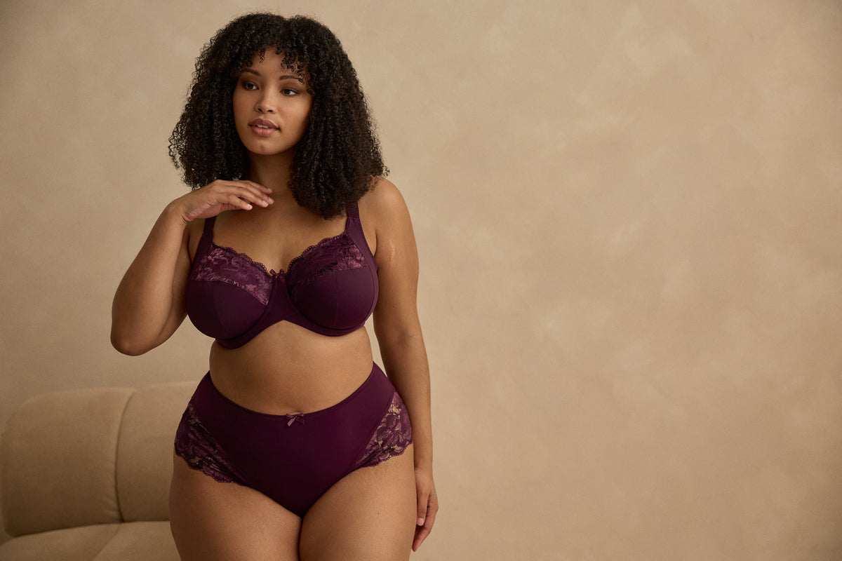 A woman modeling a matching two-piece lingerie set in deep plum purple, against a gray background
