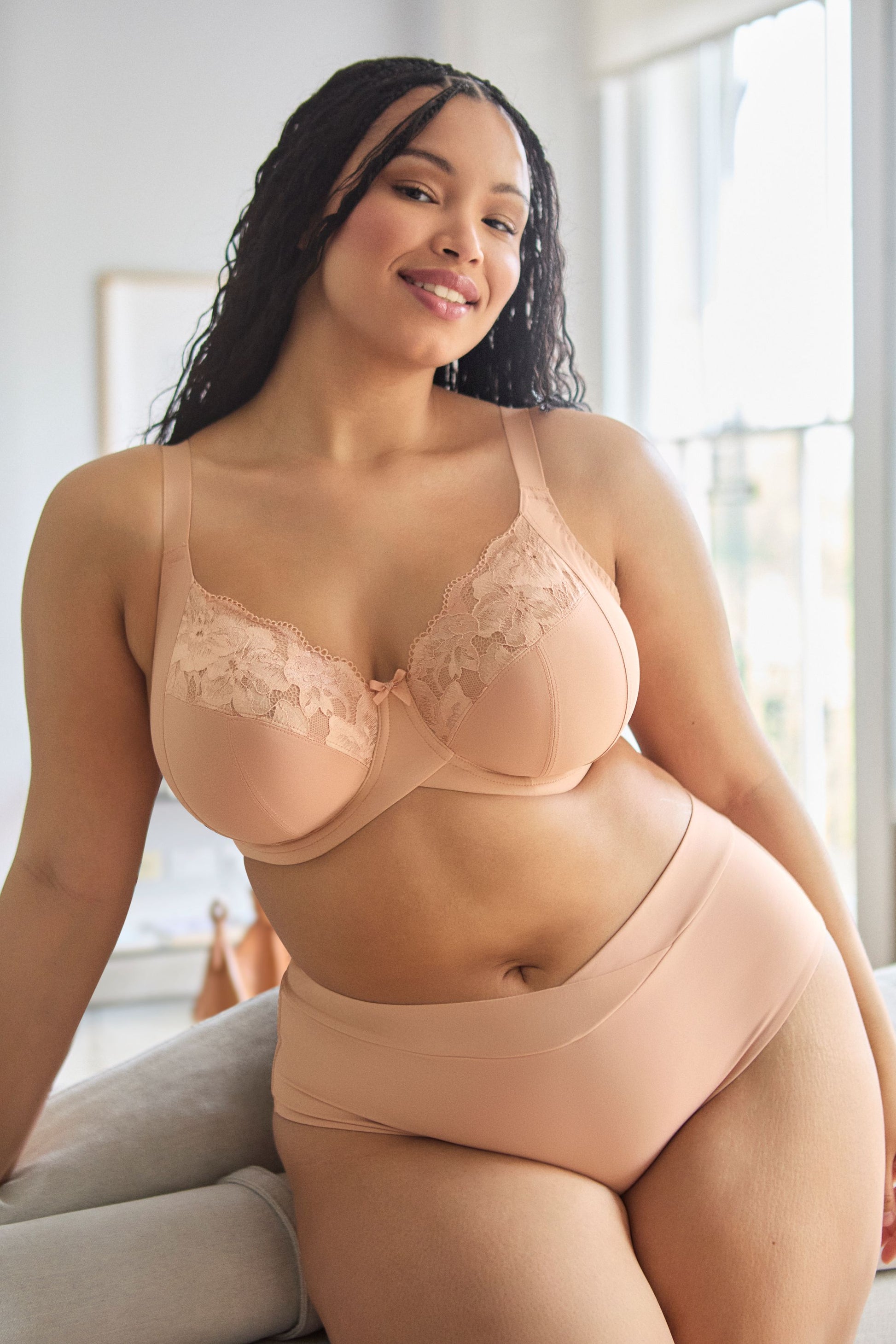 Woman wearing a beige lace bra and panties set in a bright room.