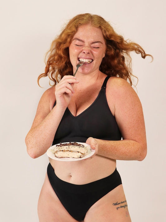 Woman in black lingerie eating dessert with a spoon against a plain background